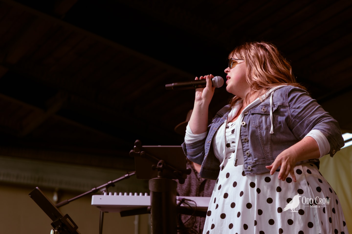 Female vocalist in polka-dot dress and denim jacket singing into microphone on outdoor stage.