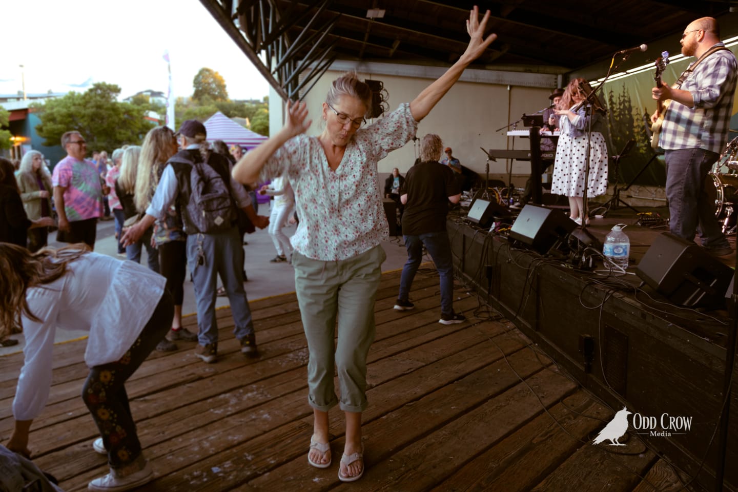 Woman dances joyfully with arms raised on wooden pier stage at outdoor summer concert.