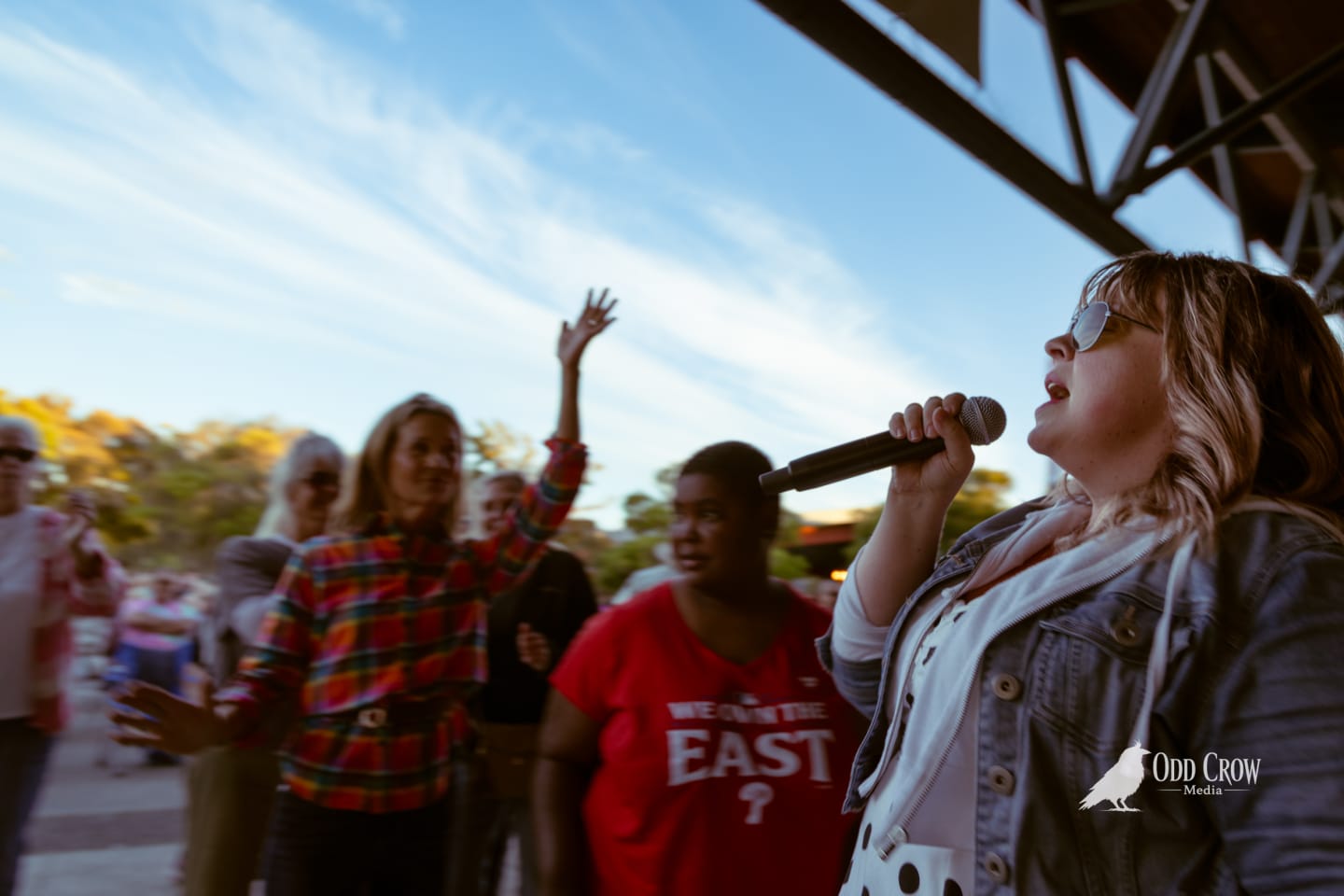 Singer performing with microphone outdoors at sunset, crowd dancing behind her on the pier.