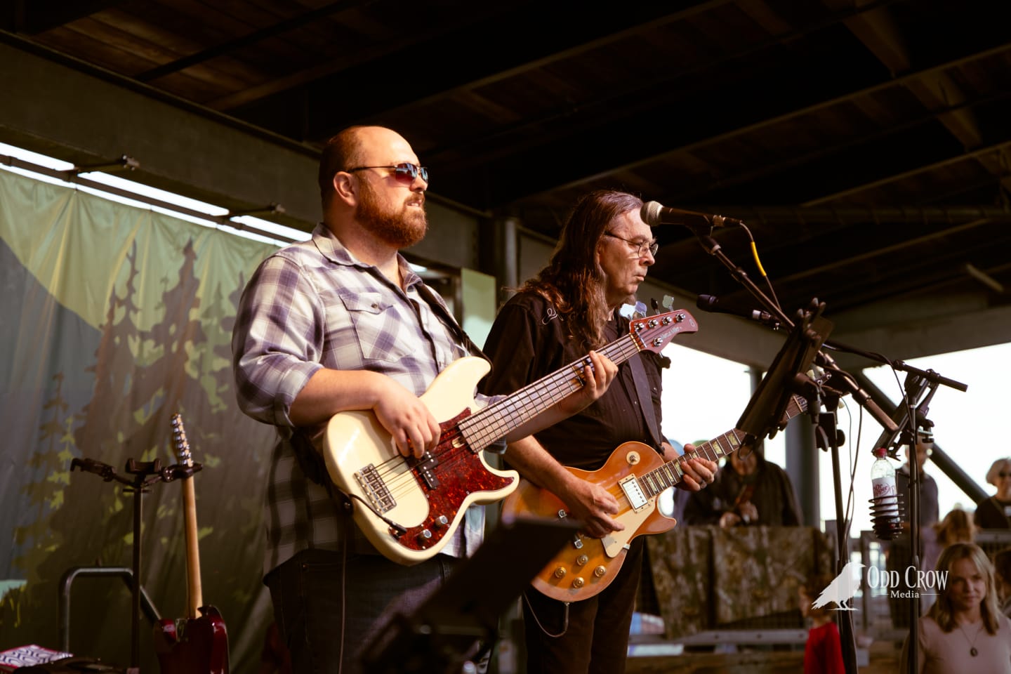 Two guitarists performing on an outdoor pier stage, one holding a cream Telecaster, the other a gold Les Paul.