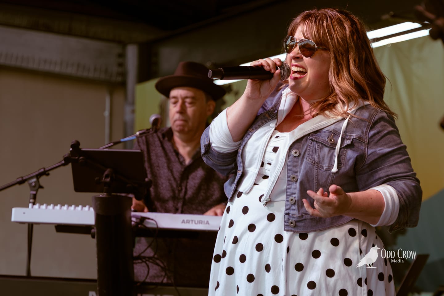 Female vocalist singing into microphone onstage, keyboardist in hat behind her, outdoor pier performance.