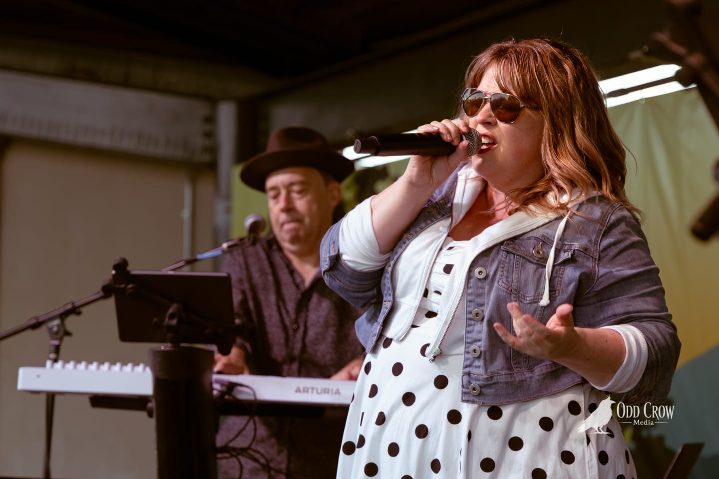 Female vocalist singing into microphone on outdoor stage, keyboardist in fedora performing behind her.