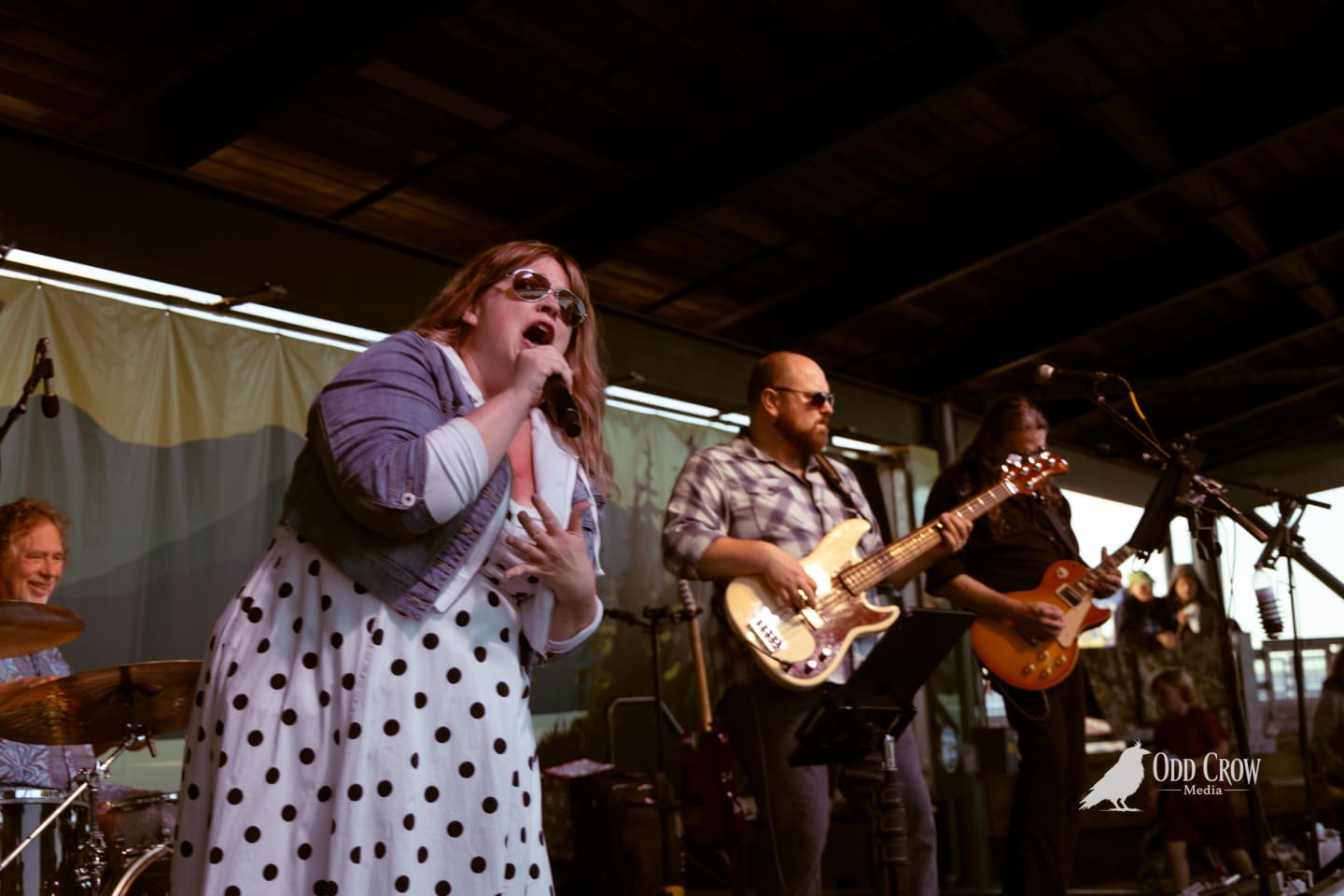 Female vocalist singing into mic with two guitarists performing on outdoor covered stage.