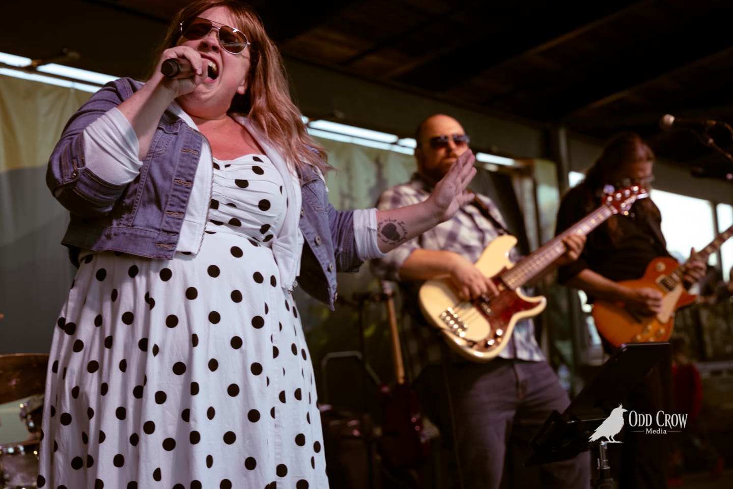 Female vocalist singing into mic, flanked by two guitarists performing on outdoor stage at waterfront venue.