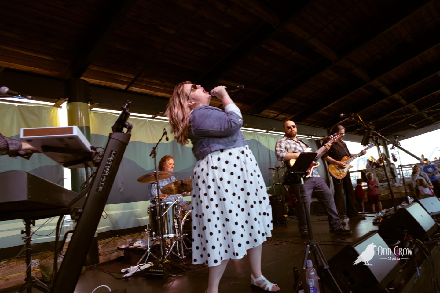 Female vocalist singing into handheld mic on outdoor pier stage with guitarist and drummer behind her.
