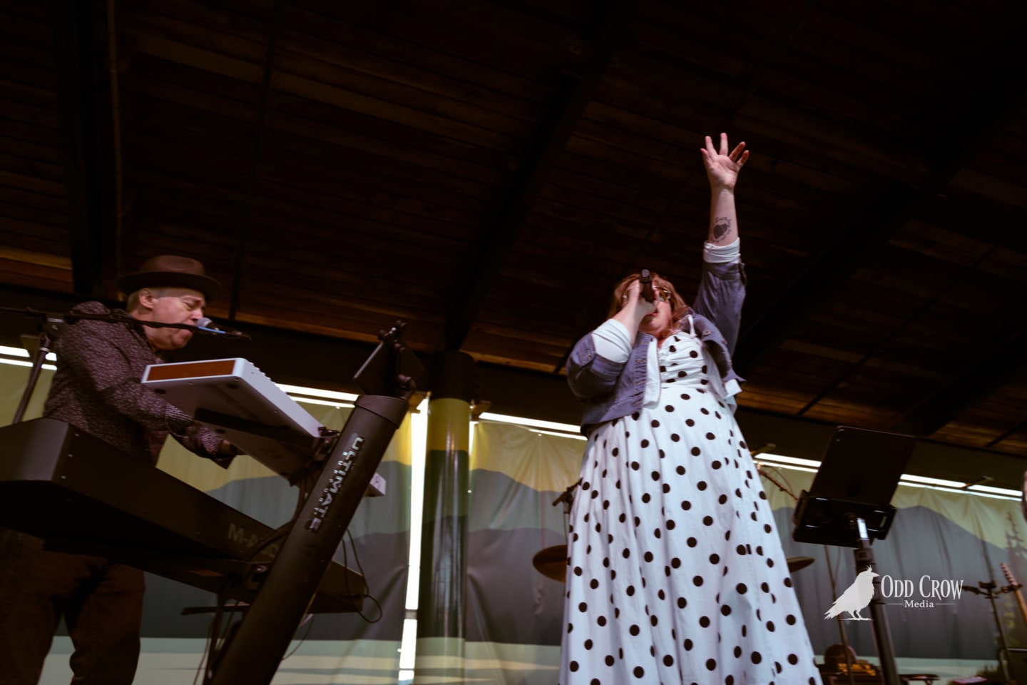 Singer in polka-dot dress raises arm mid-performance while keyboardist plays beside her on an outdoor stage.