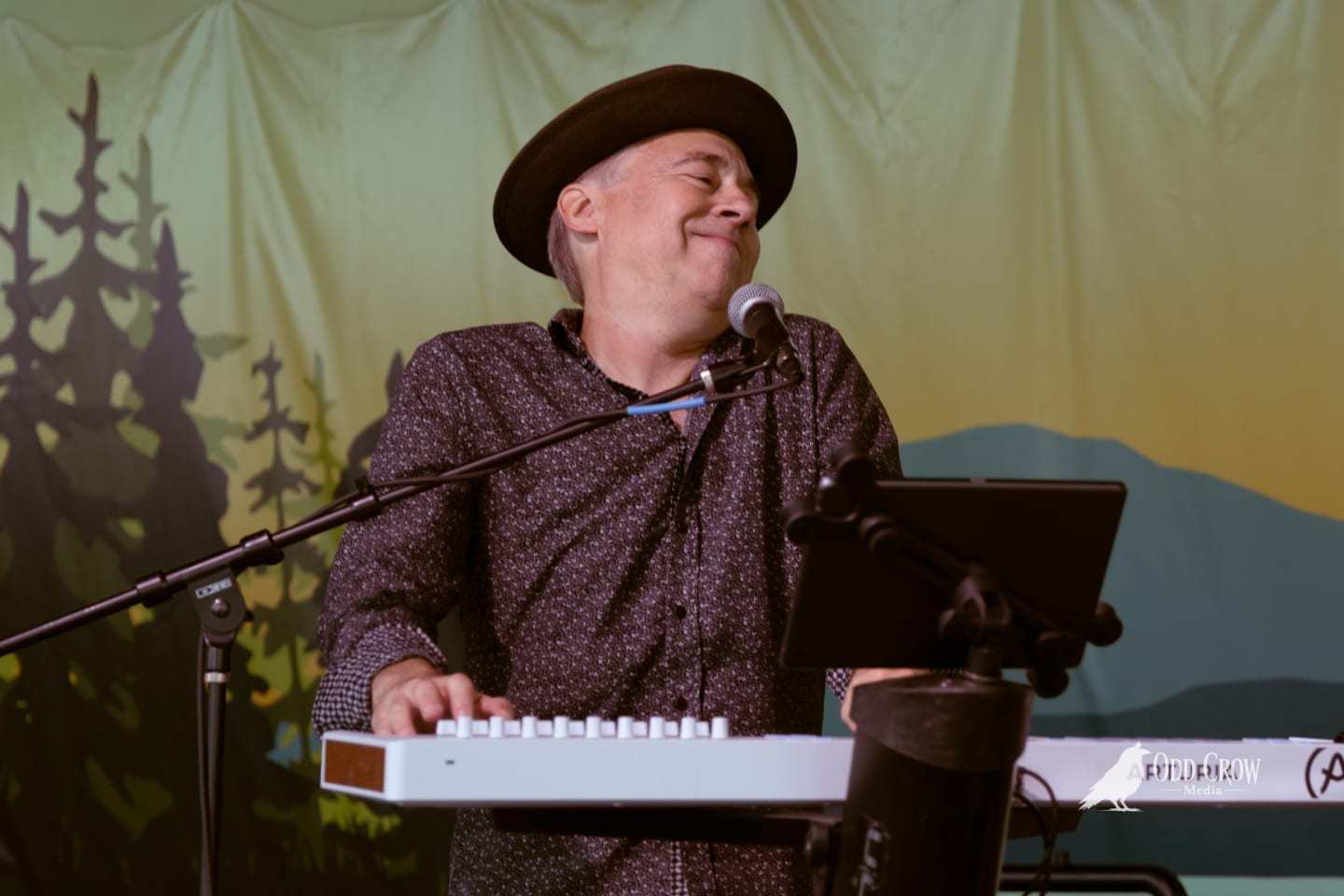 Keyboardist in a black hat smiles joyfully while performing on stage before a forest backdrop.