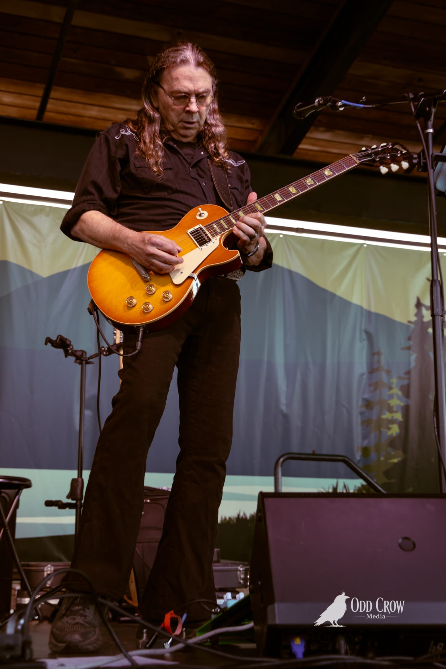 Guitarist playing a sunburst Les Paul on an outdoor stage with a painted mountain backdrop.