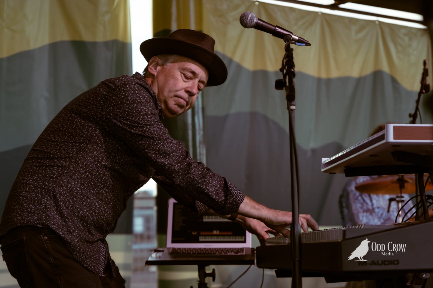 Keyboardist in a dark fedora leans intensely over his keyboard on an outdoor festival stage.