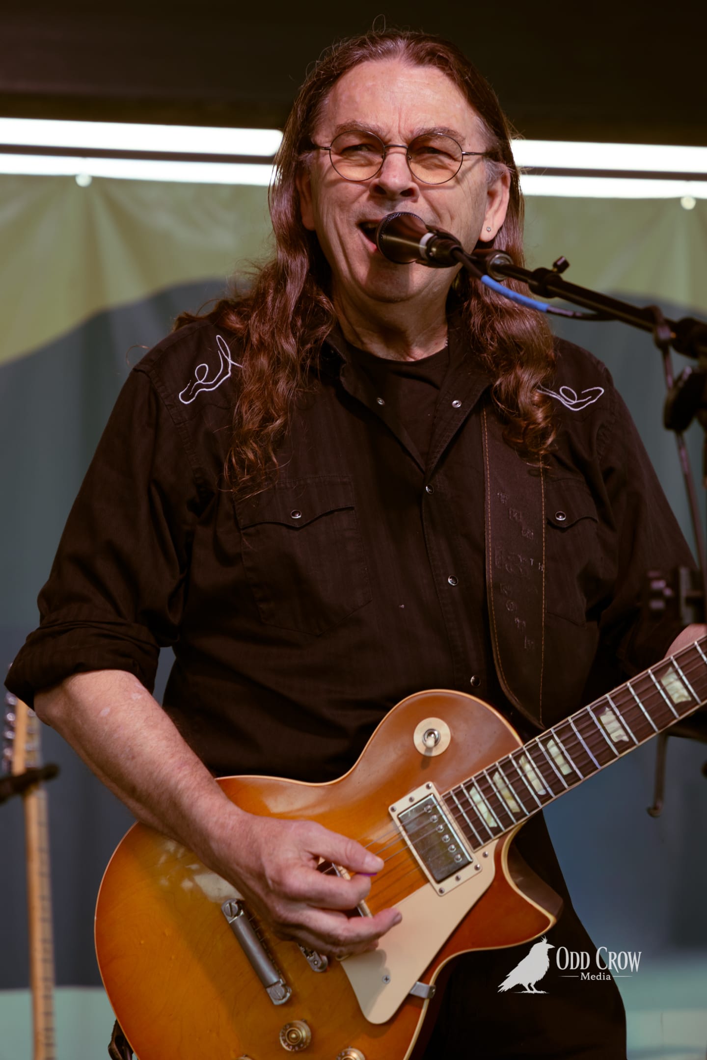 Long-haired guitarist singing into mic while playing sunburst Les Paul on outdoor stage.