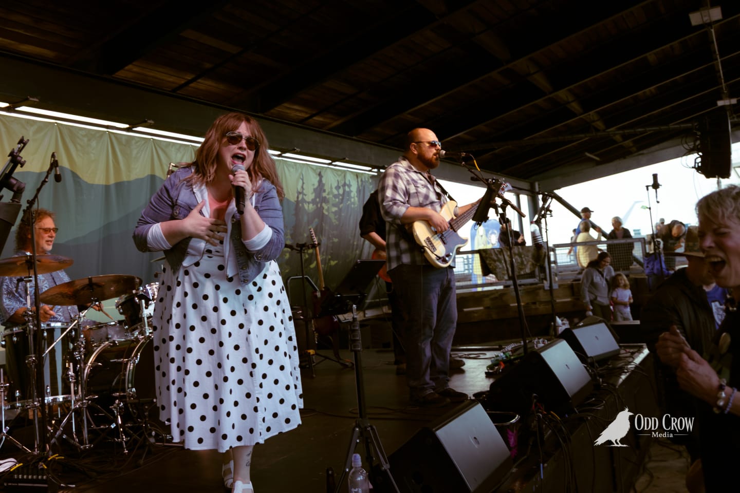 Female vocalist in polka-dot dress singing onstage at an outdoor pier venue with band behind her.