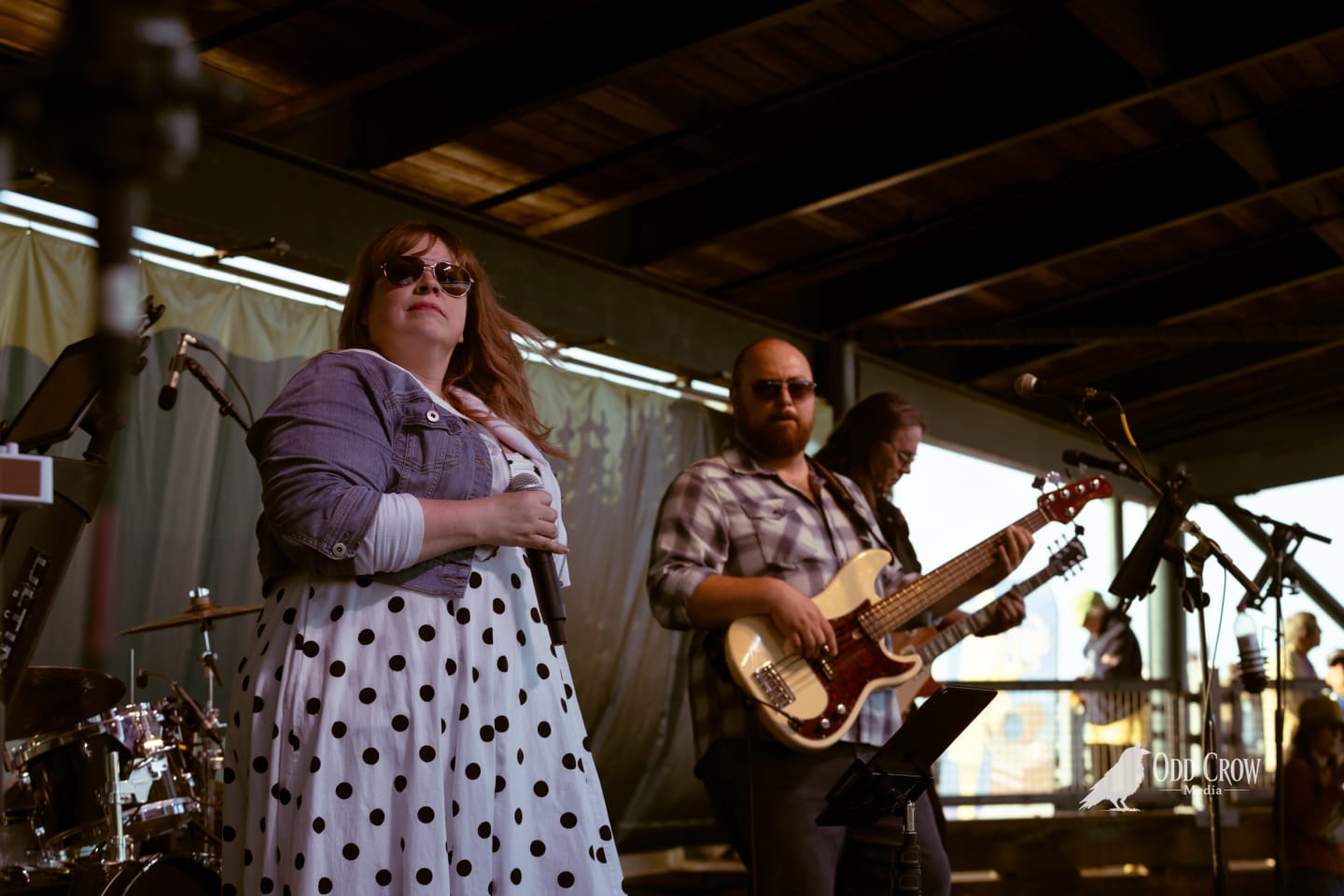 Female vocalist holding microphone performs on outdoor stage with guitarist behind her at waterfront venue.