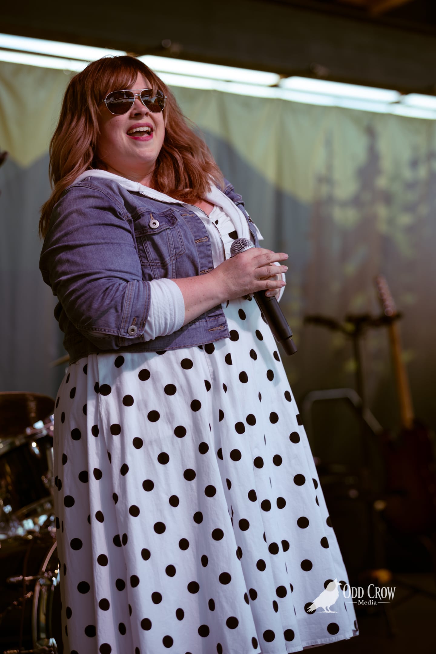 Female vocalist performing on outdoor stage in polka-dot dress and denim jacket, holding microphone.