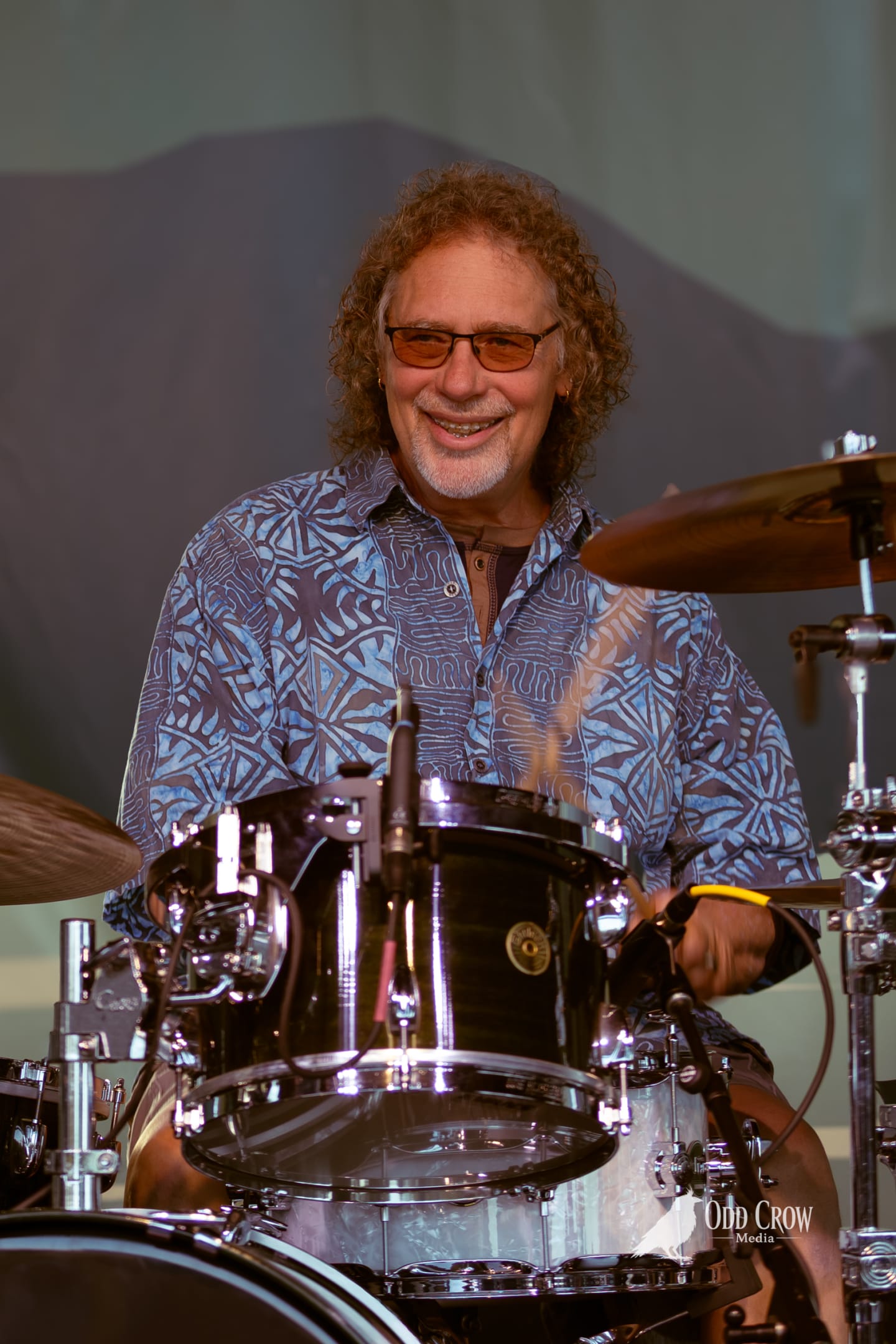 Smiling drummer with curly hair and tinted glasses plays kit on outdoor stage at sunset.