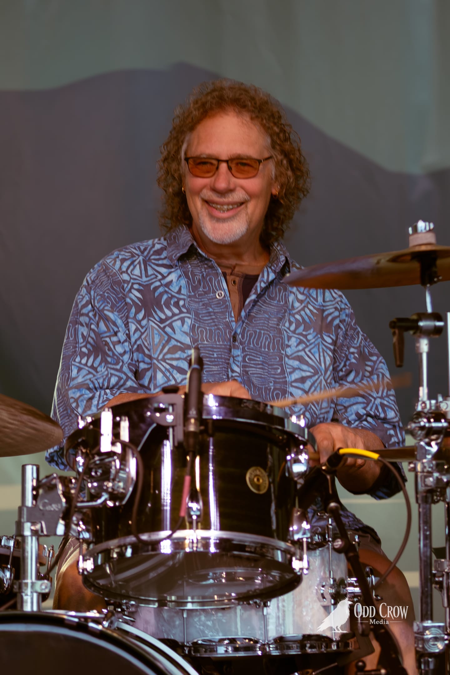 Smiling drummer with curly hair and tinted glasses plays kit on outdoor stage at dusk.