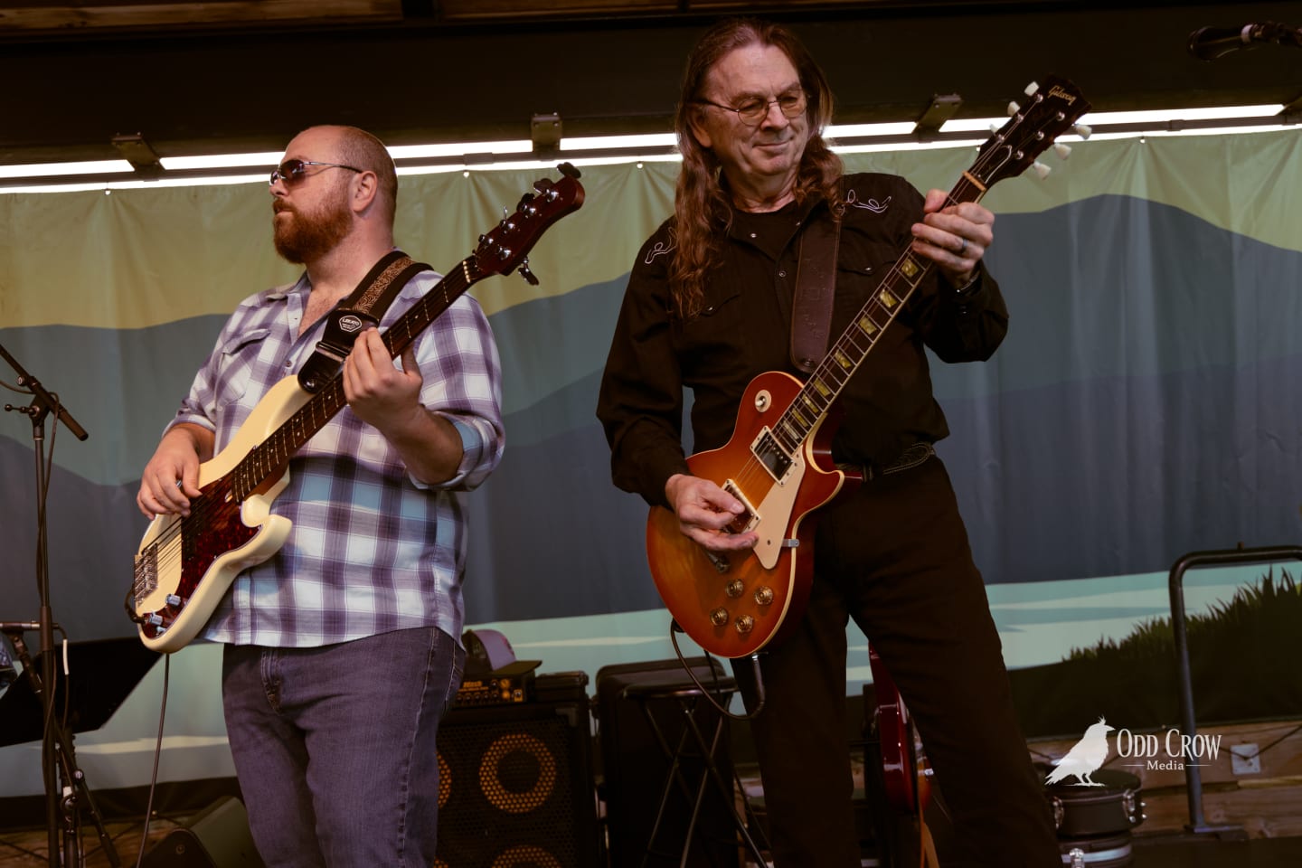 Two guitarists performing on stage, one playing a Gibson Les Paul, at an outdoor pier venue.