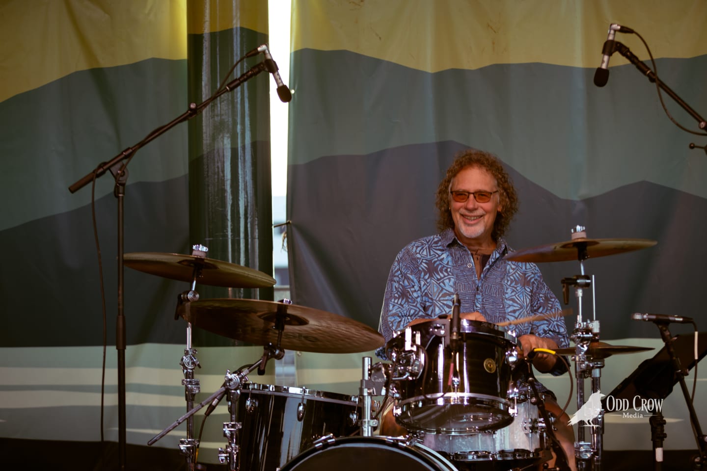 Smiling drummer with curly hair and sunglasses performs behind a full drum kit on an outdoor stage.