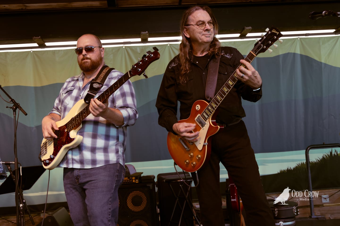 Two guitarists perform on stage at an outdoor pier venue, one playing a Gibson Les Paul.