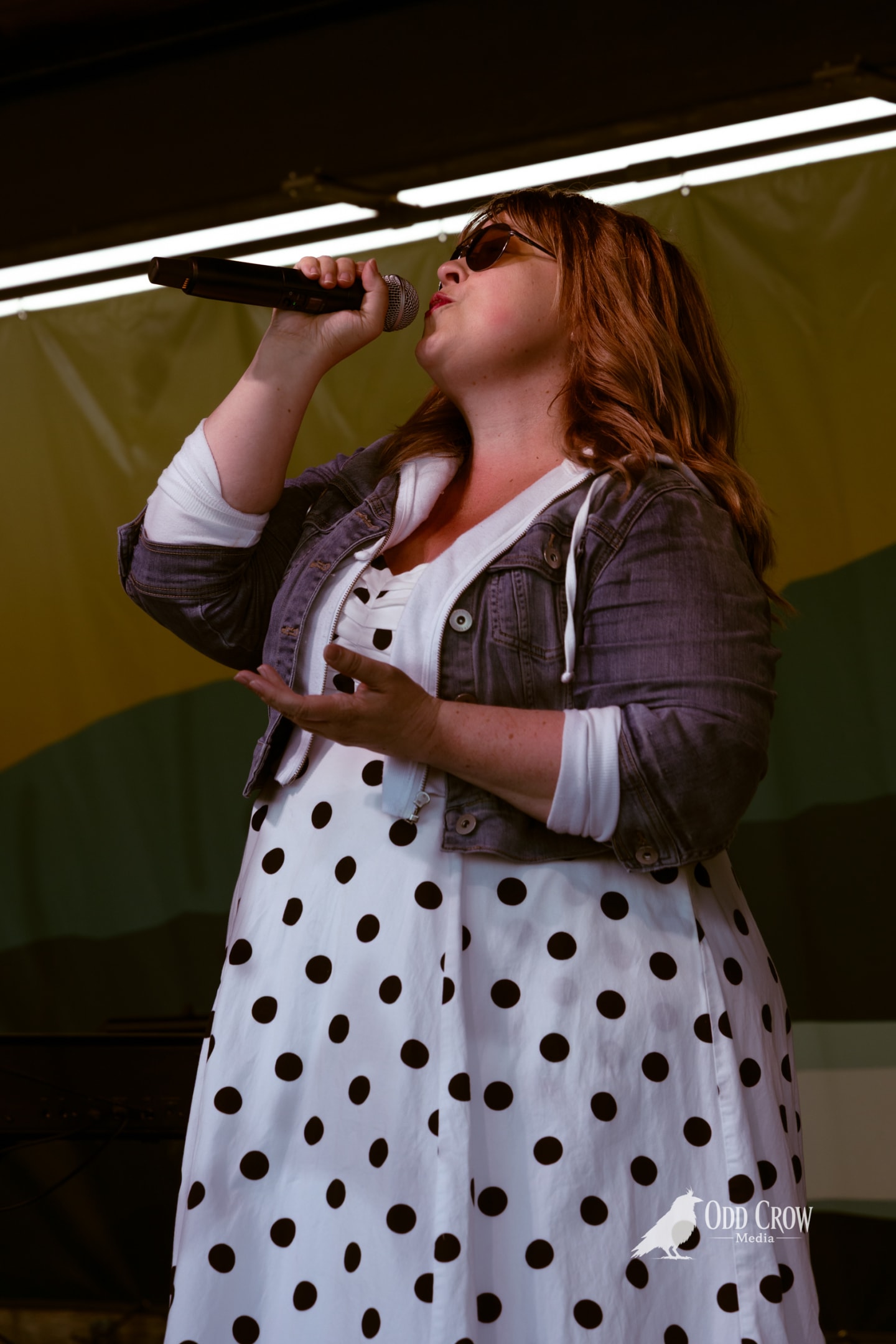 Red-haired female vocalist singing into a microphone on an outdoor stage in a polka-dot dress.