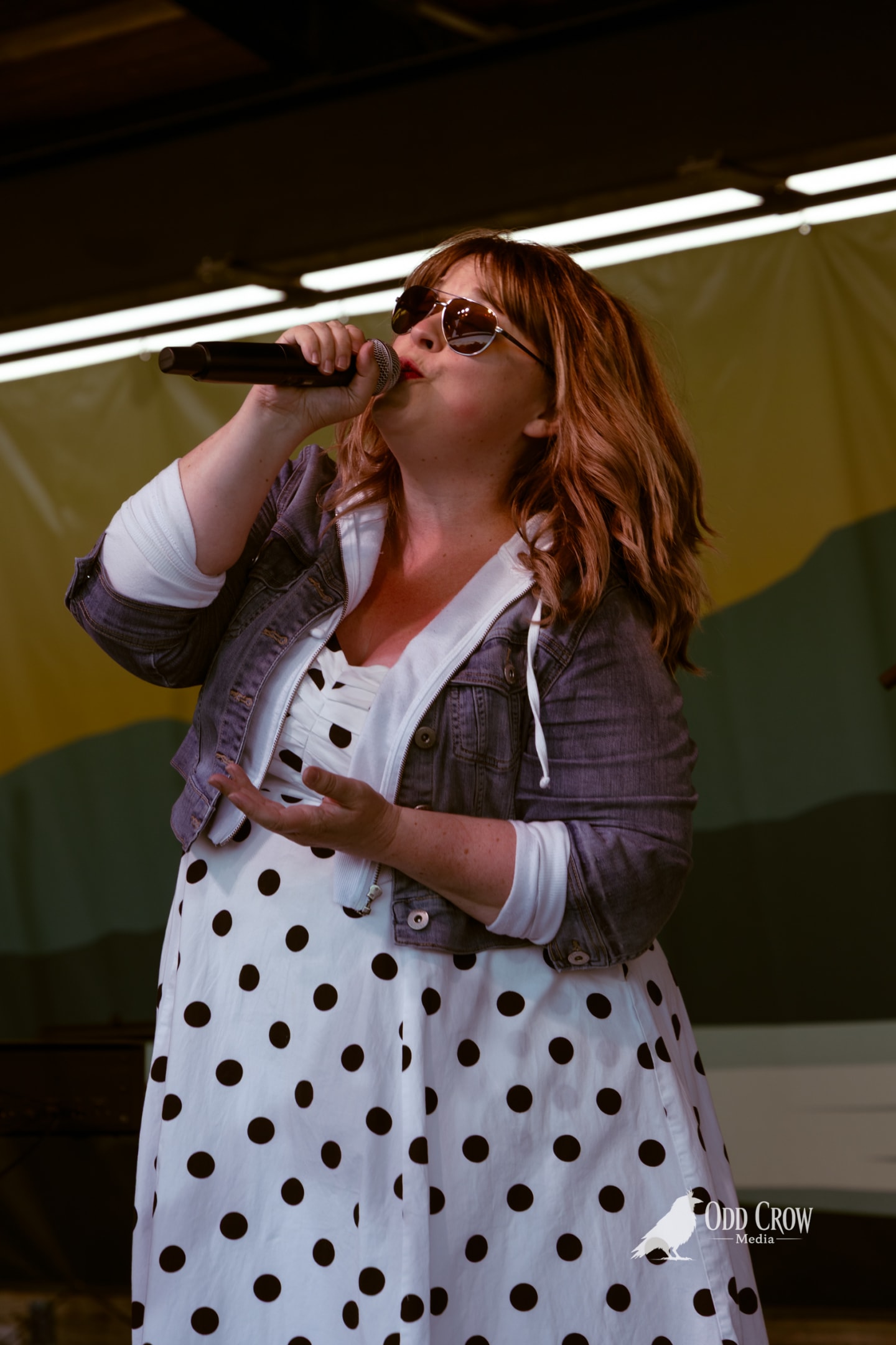 Female singer performing outdoors on stage, holding microphone, wearing polka dot dress and denim jacket.