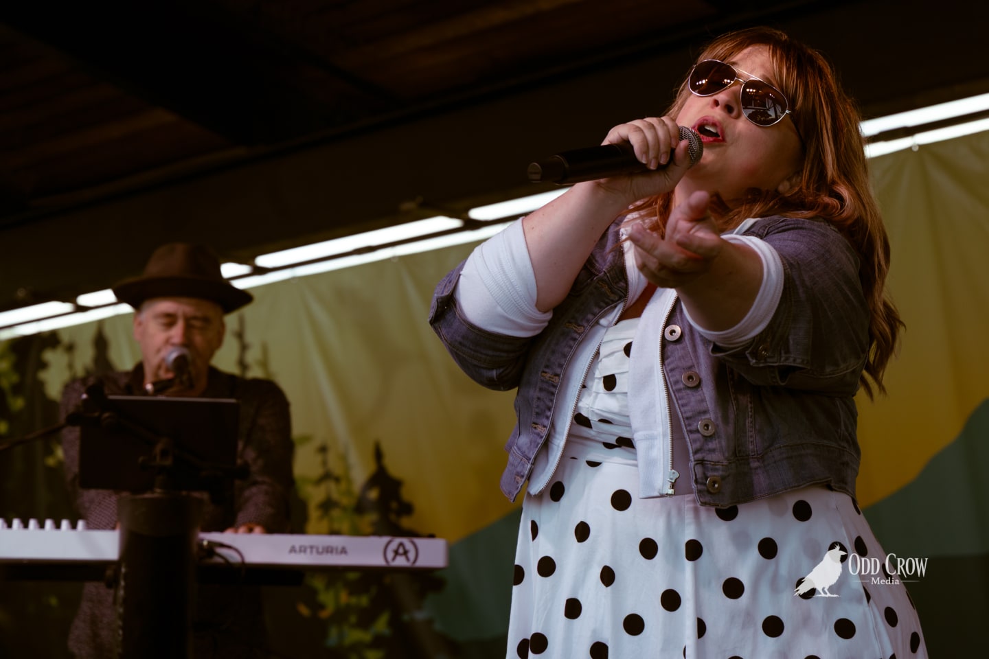 Female vocalist in polka dot dress and sunglasses performs on outdoor stage while keyboardist plays behind her.