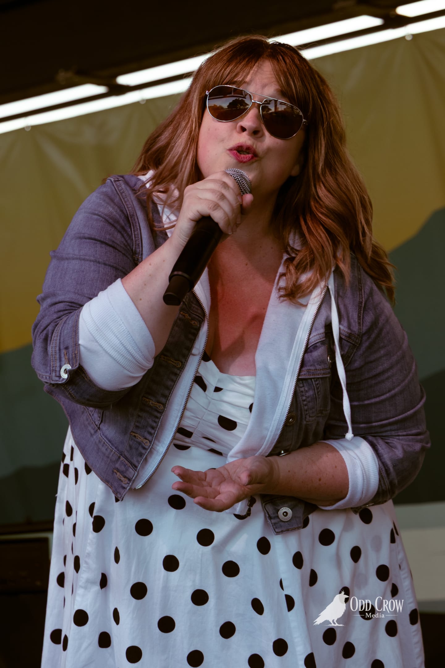 Female singer performing outdoors on stage, holding microphone, wearing polka dot dress, denim jacket, and aviator sunglasses.