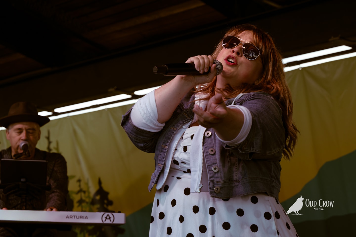 Female singer in polka-dot dress and sunglasses performs on outdoor stage with keyboardist behind her.