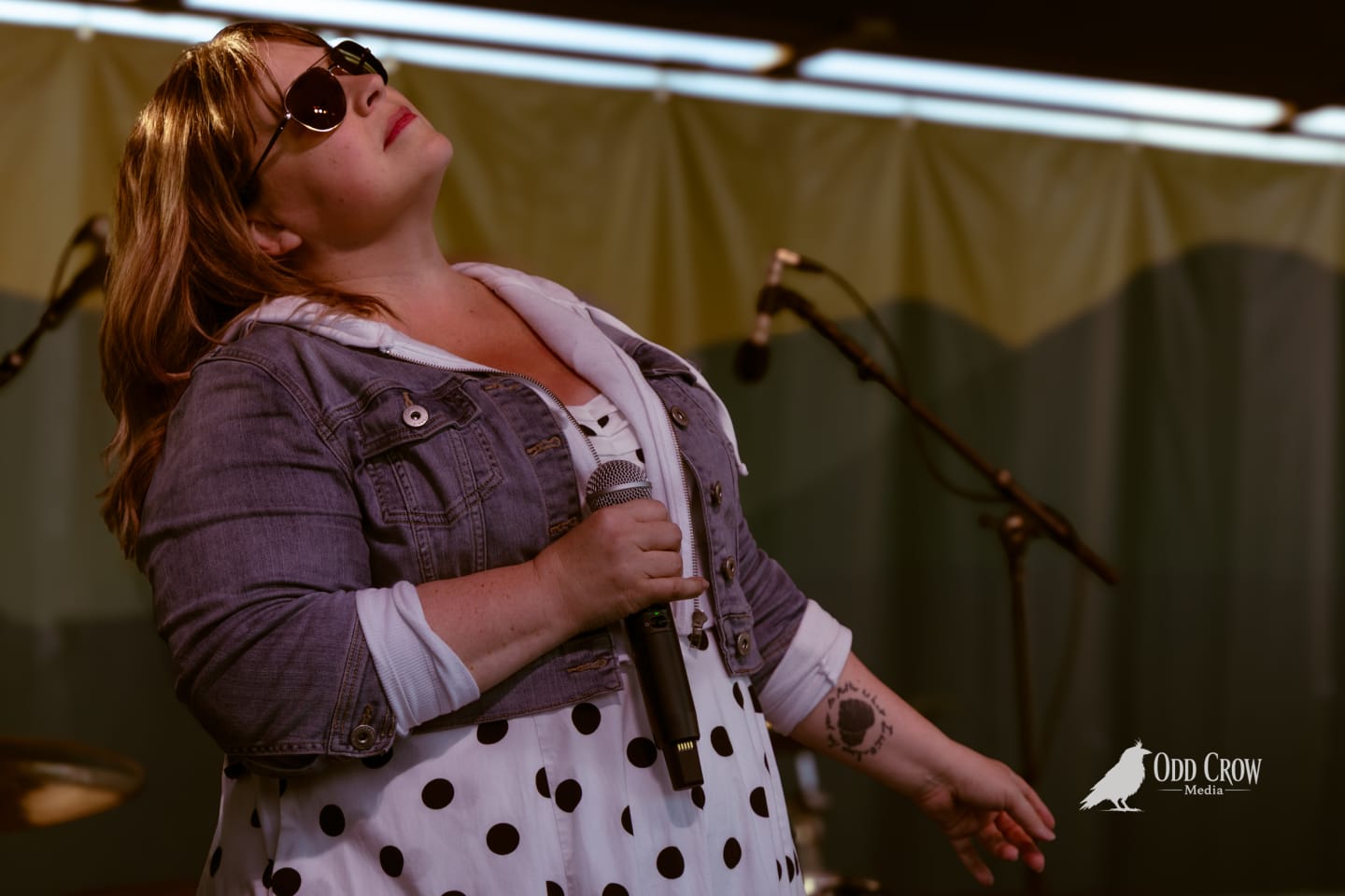 Female vocalist in sunglasses and denim jacket holds microphone onstage at an outdoor pier concert.