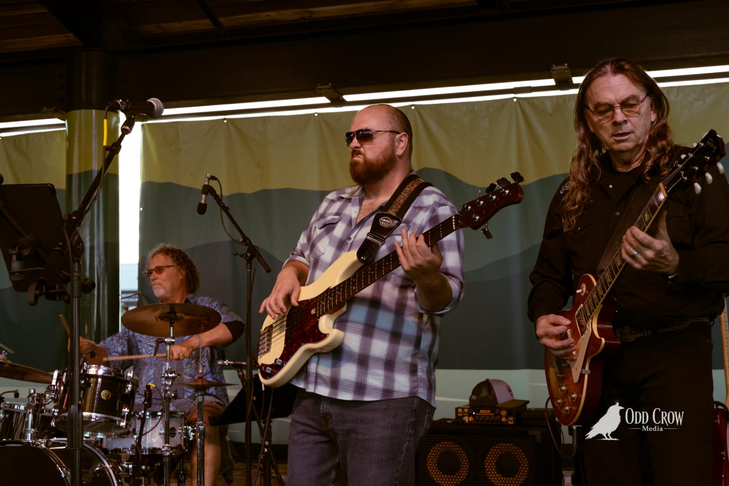 Three musicians perform live on an outdoor stage, two playing guitars and one on drums.