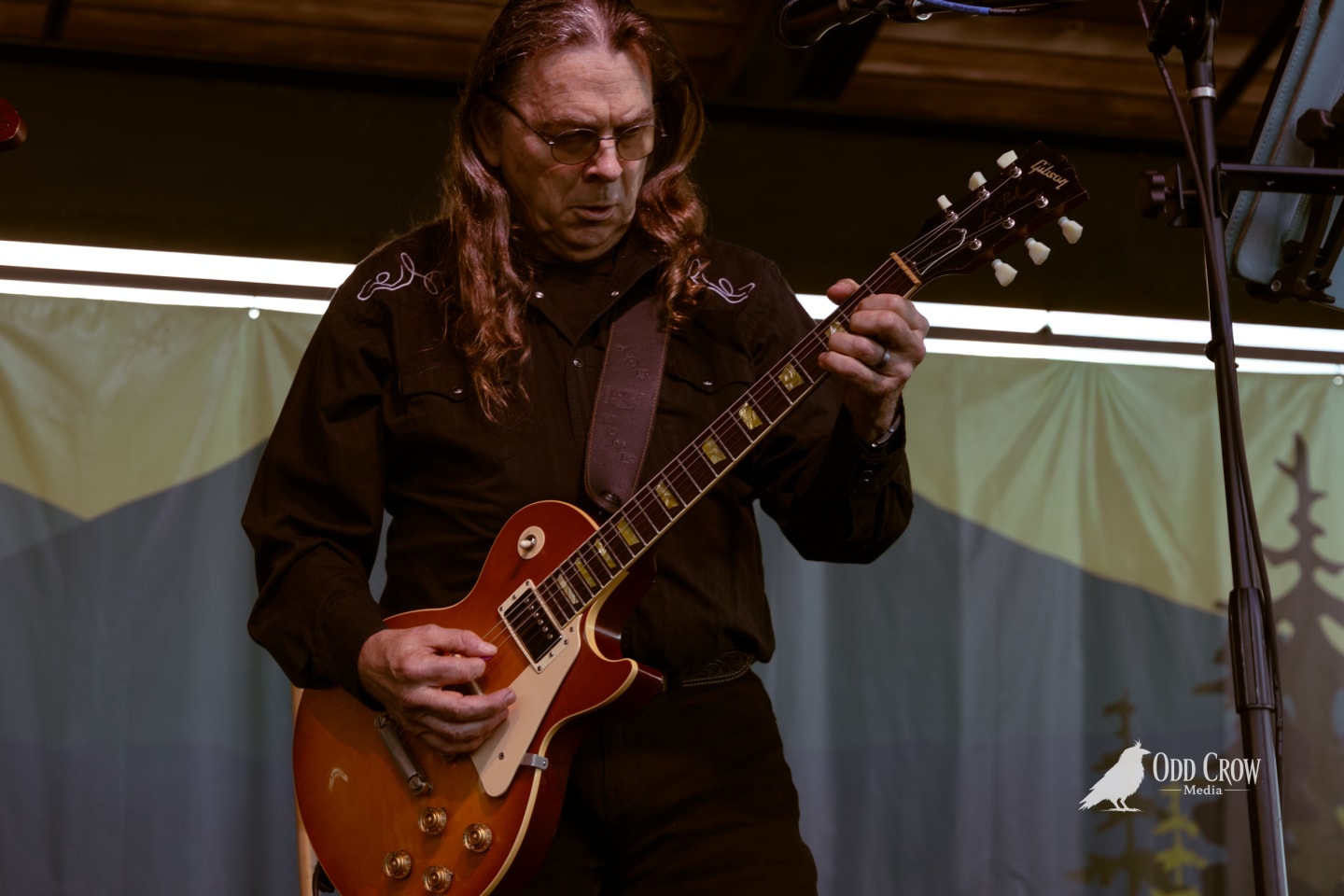 Guitarist in dark western shirt plays Gibson Les Paul on outdoor stage at waterfront concert.