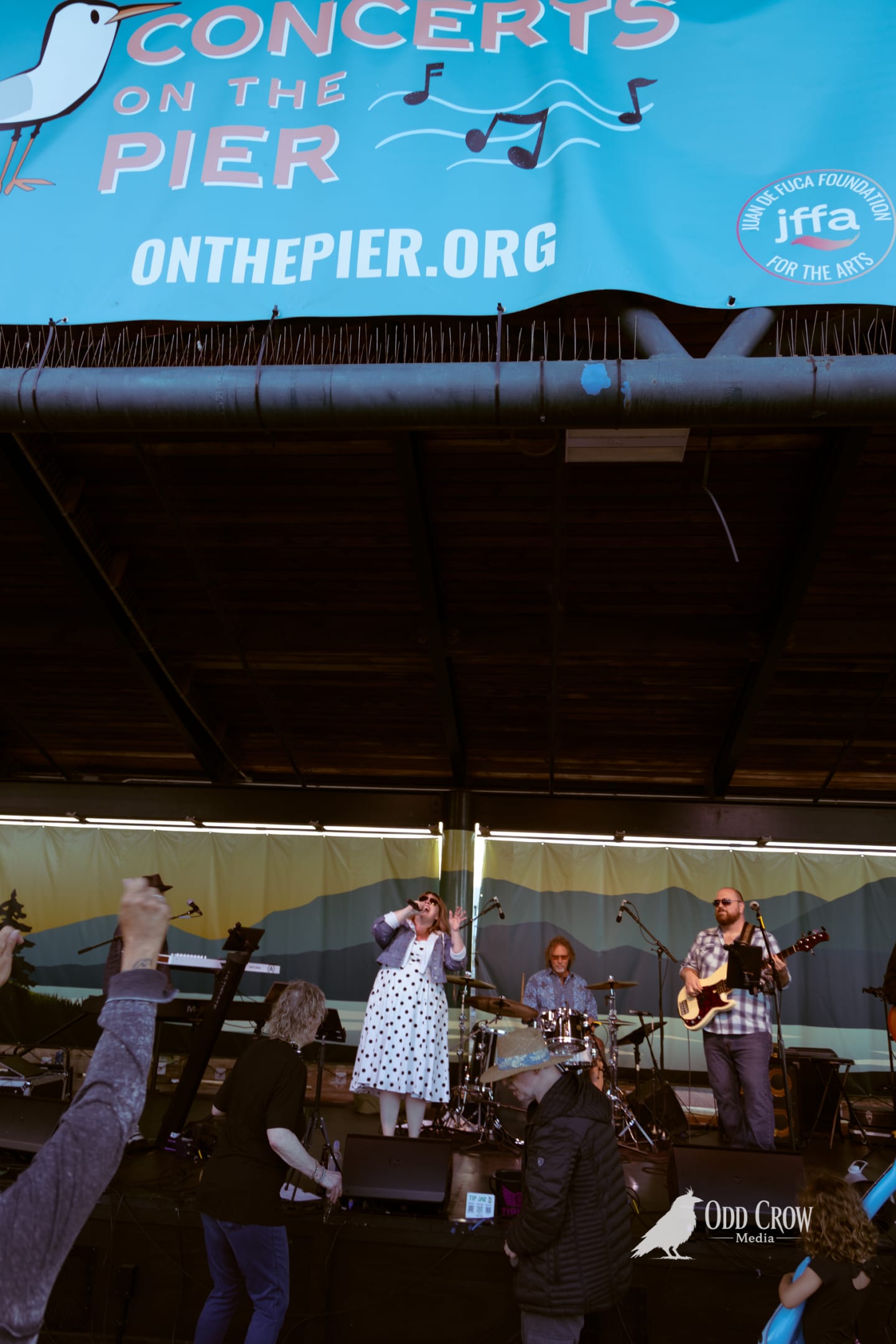 Band performs on covered pier stage beneath Concerts on the Pier banner as dancer raises arms in foreground.