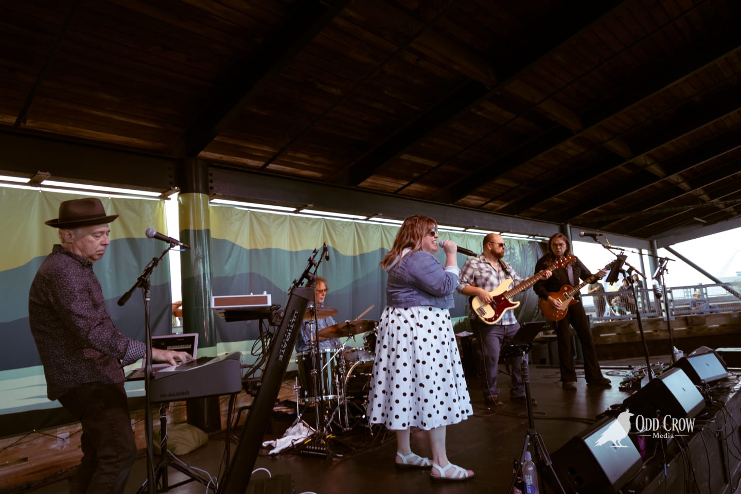 Live band performs on covered pier stage, female vocalist in polka-dot skirt singing into microphone.