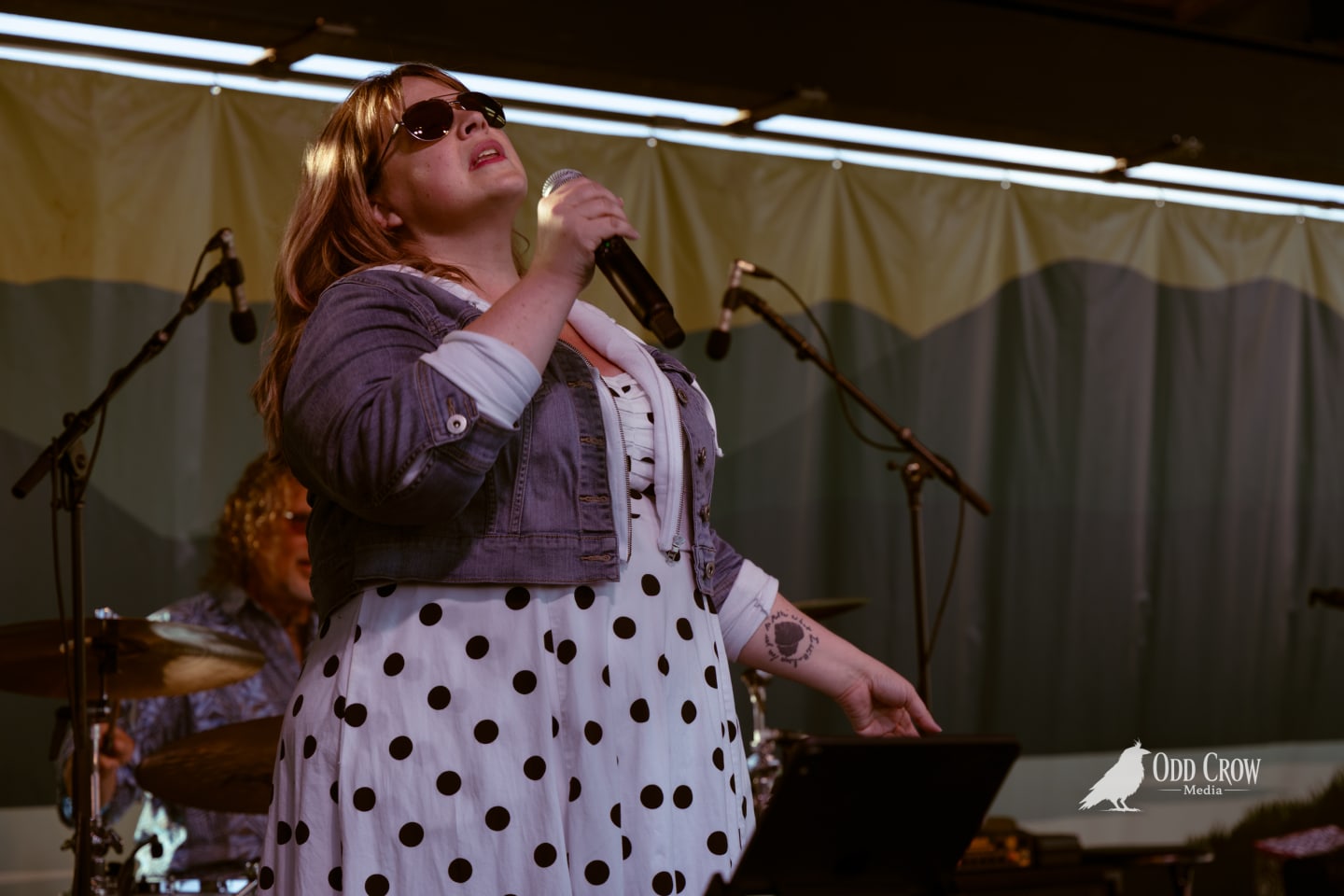 Female vocalist in polka dot dress and sunglasses singing into microphone on outdoor stage at pier concert.