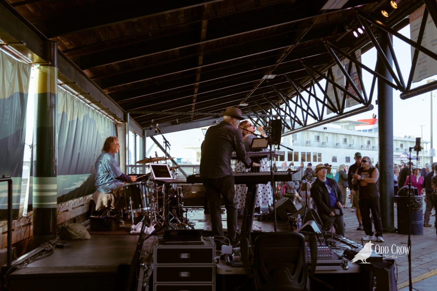 Band performing on covered pier stage at waterfront, with crowd and docked vessel visible behind them.