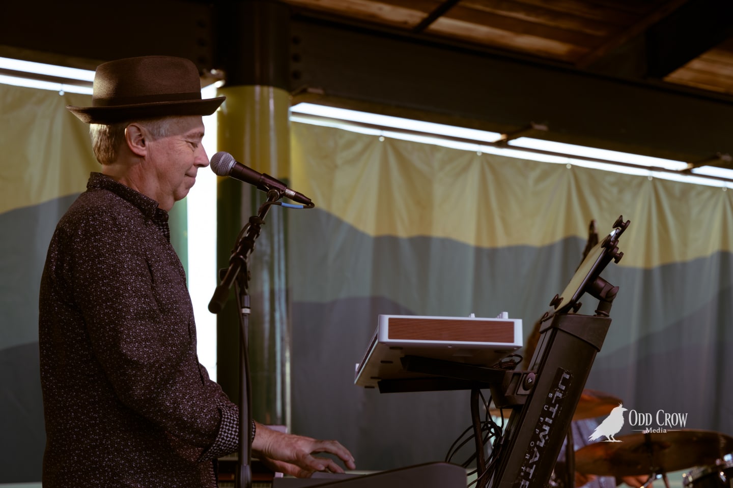 Keyboard player in a brown fedora performs at an outdoor stage with colorful striped backdrop.