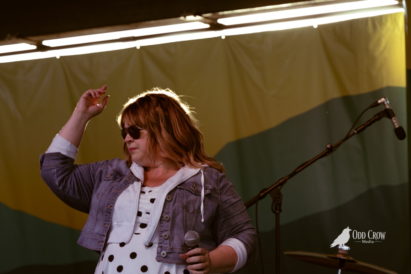 Female vocalist holding microphone on outdoor stage, one hand raised, wearing sunglasses and denim jacket.