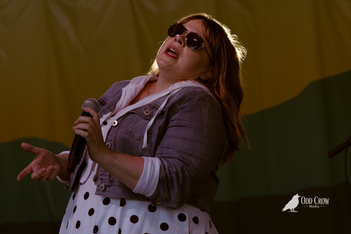 Female vocalist singing into microphone on outdoor stage, wearing sunglasses and polka-dot dress with denim jacket.