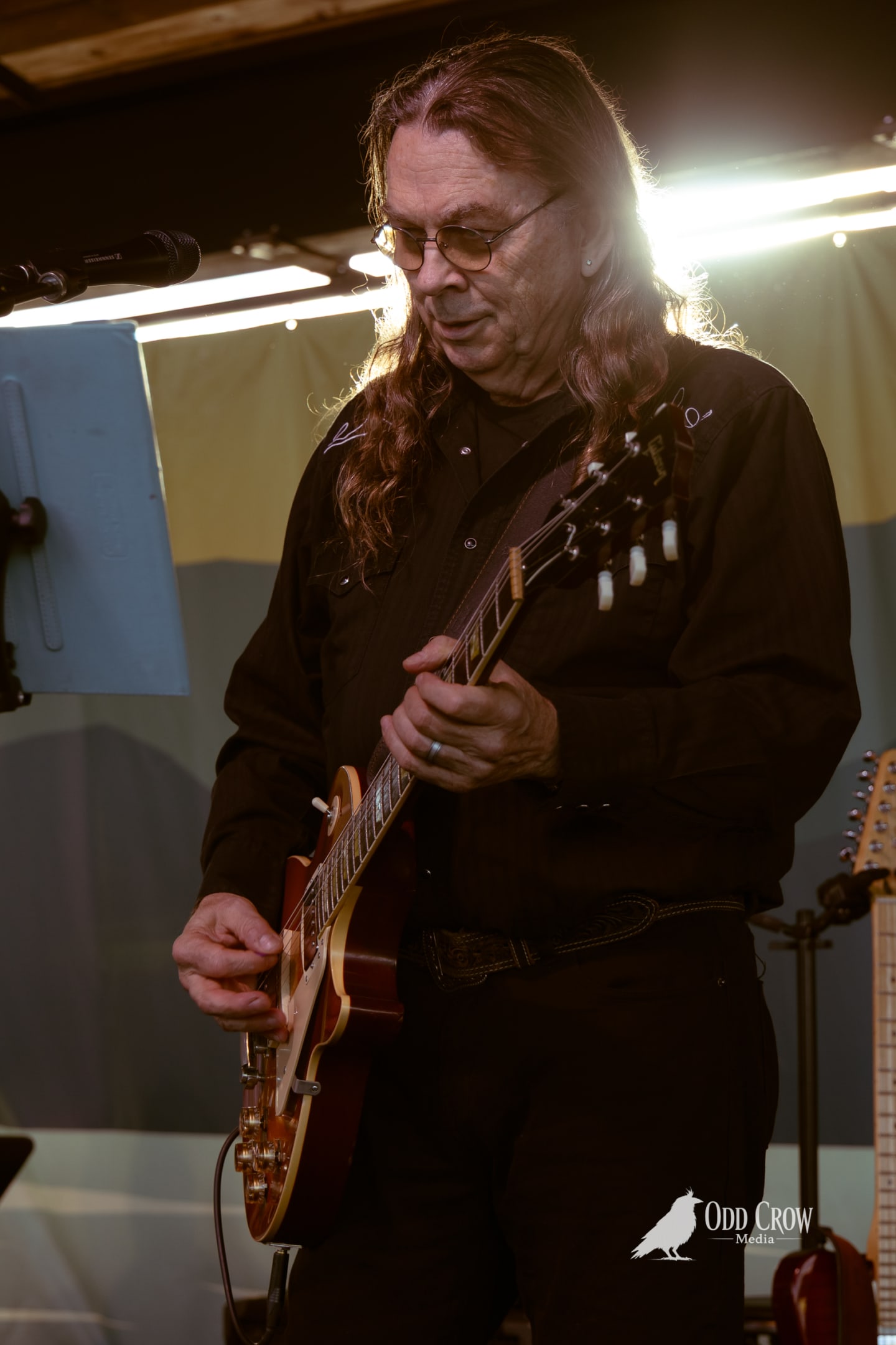 Guitarist with long hair and glasses plays electric guitar on stage under bright lights.