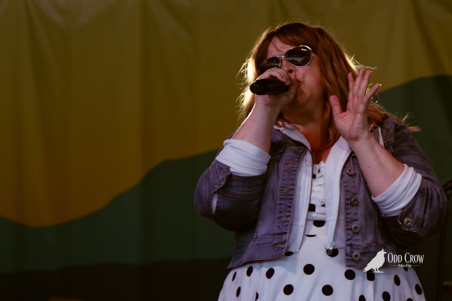 Female singer performing on outdoor stage, holding microphone, wearing sunglasses, polka dot dress, and denim jacket.