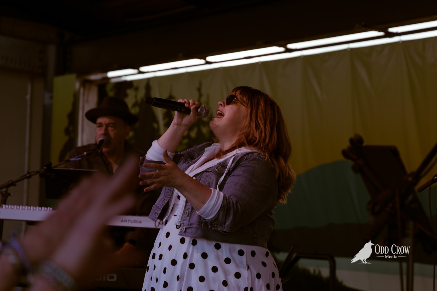 Female vocalist singing into handheld mic on outdoor stage, wearing polka-dot dress and sunglasses, keyboardist visible behind her.