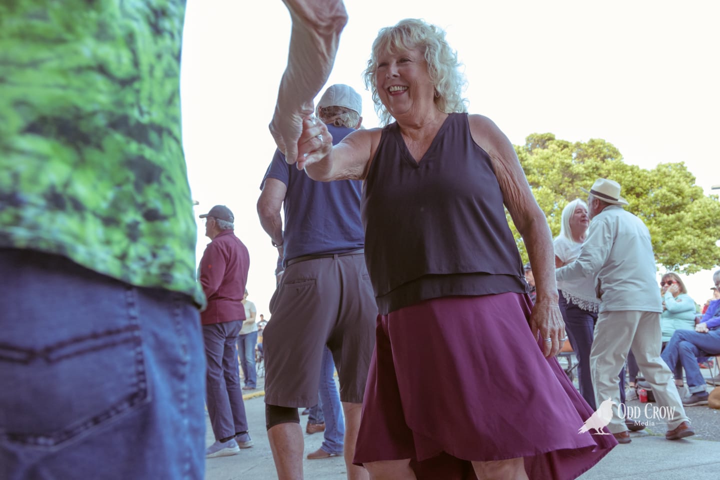Smiling woman in purple skirt dances with a partner at an outdoor pier event.