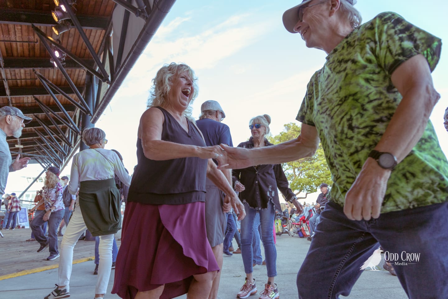 Older couple laughing and dancing together under a covered outdoor pier stage at sunset.