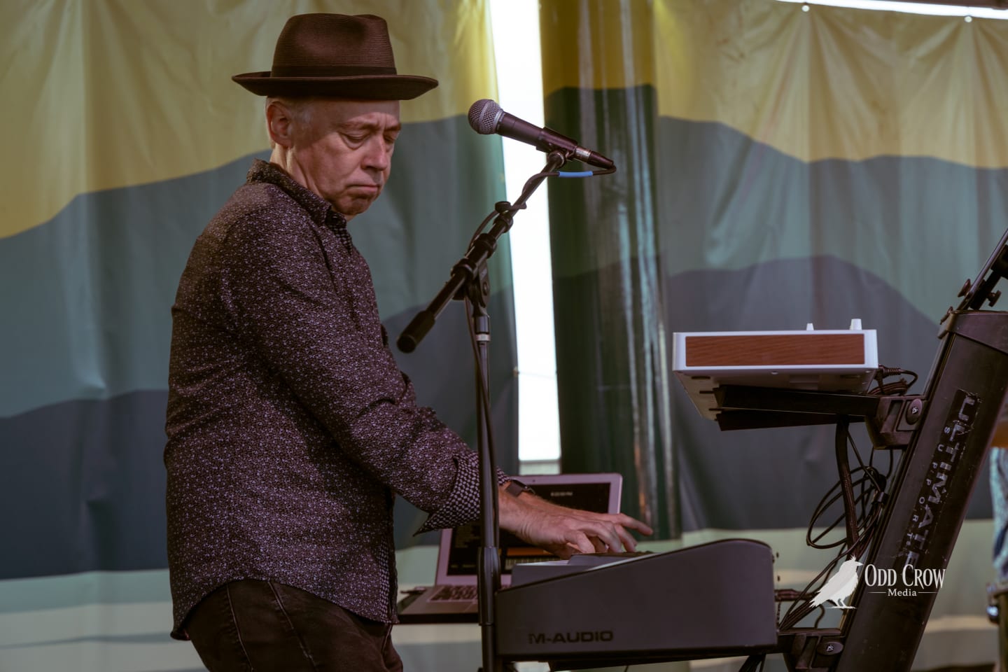 Keyboardist in a wide-brim hat plays an M-Audio keyboard on an outdoor stage.