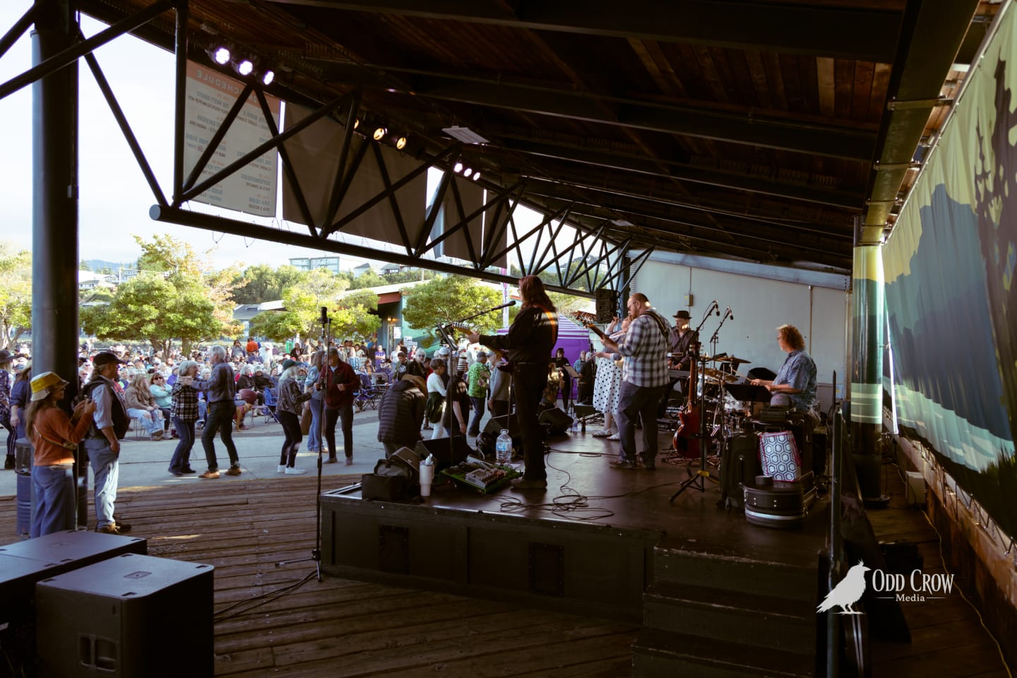 Band performs on covered outdoor stage at sunset as crowd dances and gathers at the pier.