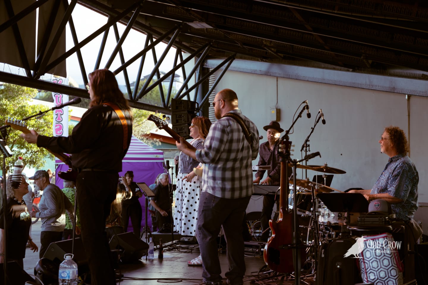 Band performing on covered outdoor stage at dusk before a festival crowd.