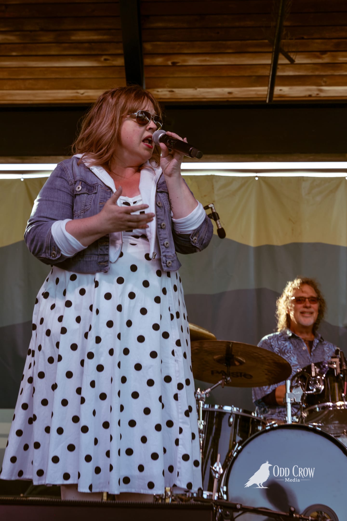 Female vocalist in polka dot dress and sunglasses singing onstage with drummer visible behind her.