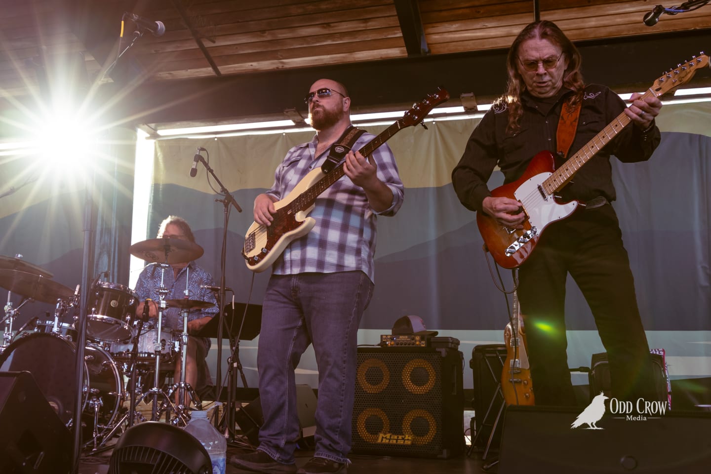 Two guitarists perform on an outdoor stage at sunset, with a drummer visible behind them.