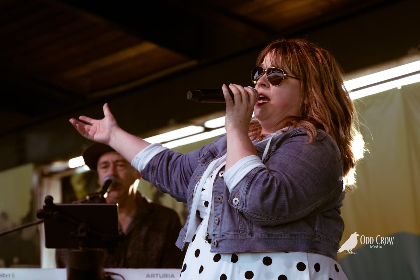 Female vocalist singing into microphone on outdoor stage, gesturing expressively, wearing sunglasses and denim jacket over polka-dot dress.