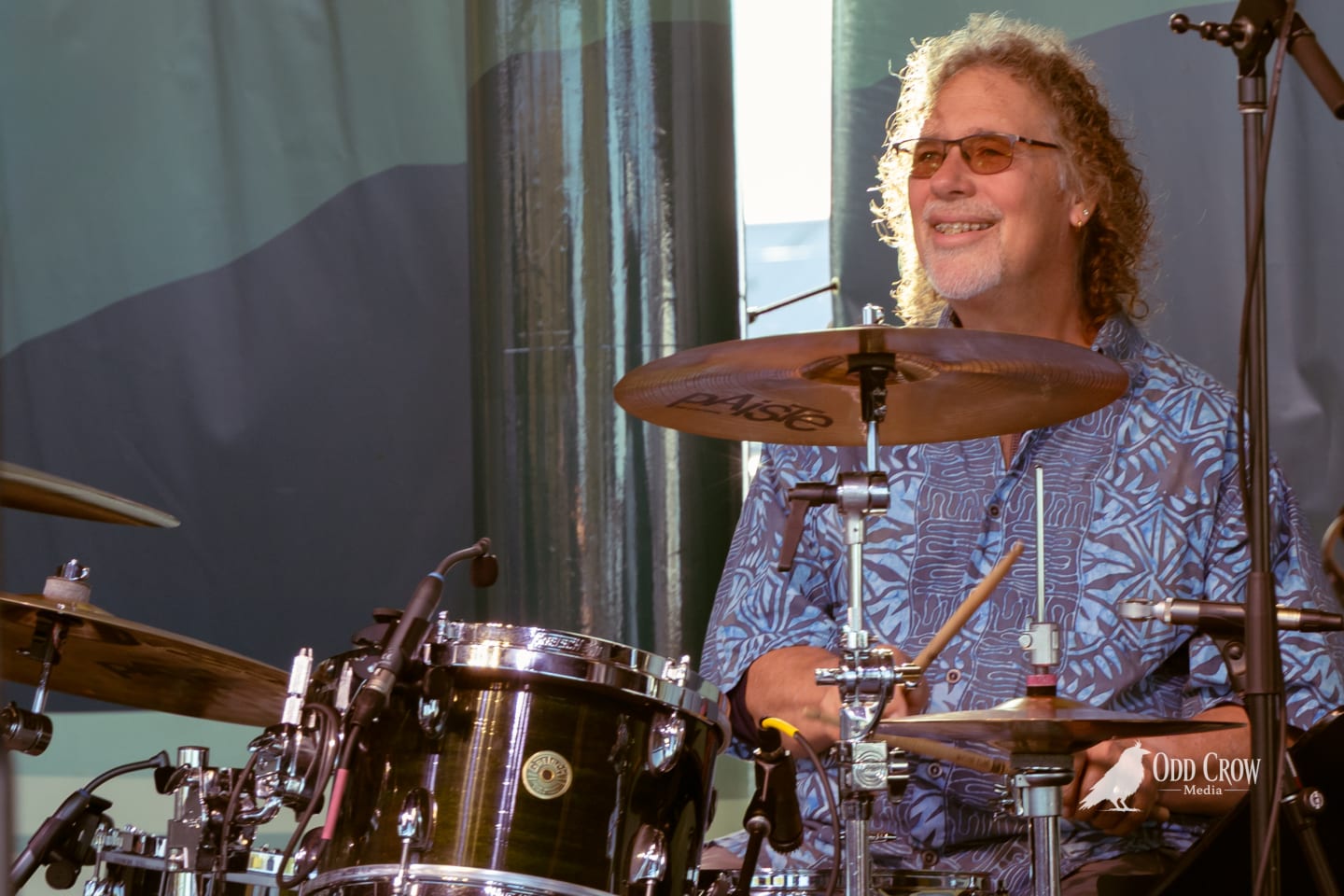 Smiling drummer with curly hair and sunglasses plays Paiste cymbals on an outdoor stage.