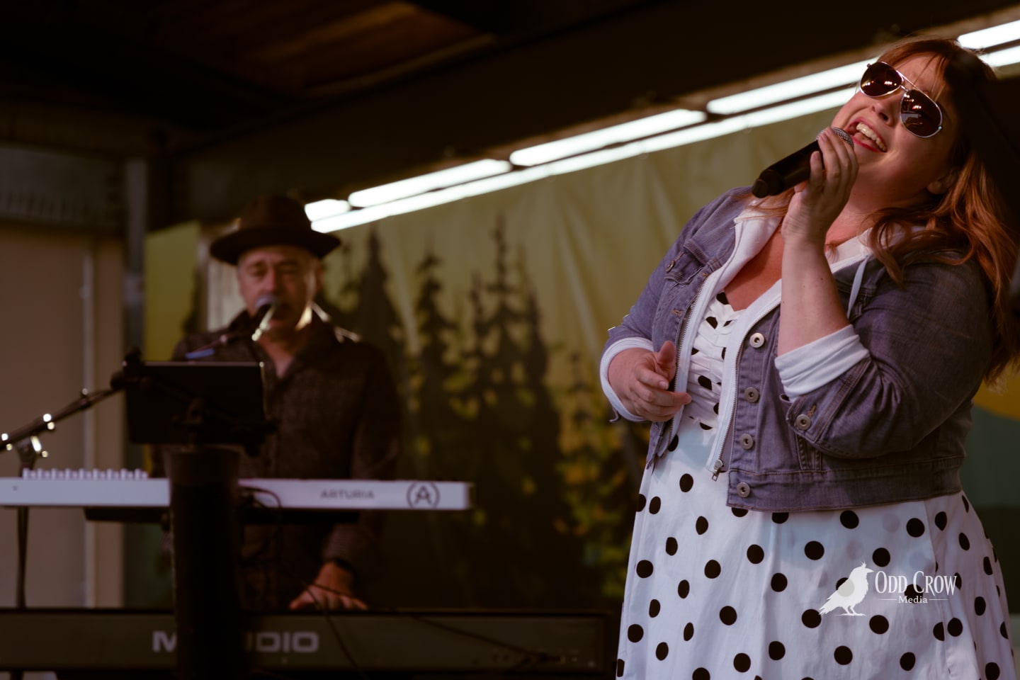 Female vocalist in polka dot dress and sunglasses singing onstage with keyboardist at outdoor summer concert.